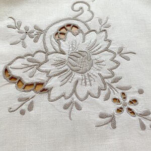 May include: A cream-colored textile featuring intricate beige thread embroidery in a floral pattern. The design includes a large central flower with detailed petals and smaller surrounding blossoms, accented by cutwork sections that reveal the surface.