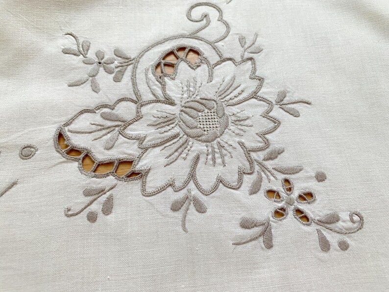 May include: A cream-colored textile featuring intricate beige thread embroidery in a floral pattern. The design includes a large central flower with detailed petals and smaller surrounding blossoms, accented by cutwork sections that reveal the surface.