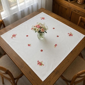Pode incluir: Uma toalha de mesa quadrada branca com motivos florais bordados em tons de rosa, vermelho e verde, espalhados pelo tecido. Um vaso de vidro transparente contendo um buquê de flores rosas e brancas está no centro da toalha.