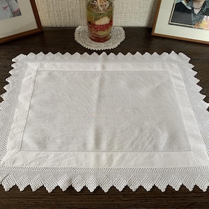 May include: A white rectangular placemat with a crocheted lace border. The placemat has a simple design with a stitched border. The lace border features a scalloped edge. The placemat is placed on a dark wooden surface.