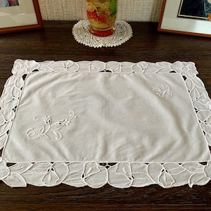 Peut inclure: Nappe rectangulaire blanche brodée avec une bordure florale et de feuilles. Le centre de la nappe présente des motifs floraux brodés. La nappe est posée sur une surface en bois brun foncé.