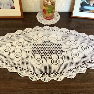 May include: Oval, white crocheted lace table runner with a floral design. The runner is placed on a dark brown wooden table. The edges have a scalloped design. Two framed photos and a glass with a coaster are also on the table.