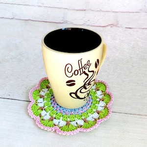 May include: A crocheted coaster in shades of pink, green, and white with a beige coffee mug with the word "Coffee" printed on it.