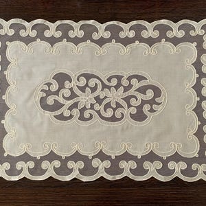 May include: Ivory-colored rectangular doily with a delicate, openwork design. The center features a floral and scroll motif, framed by a scalloped border. This decorative piece adds a touch of elegance to any table setting.