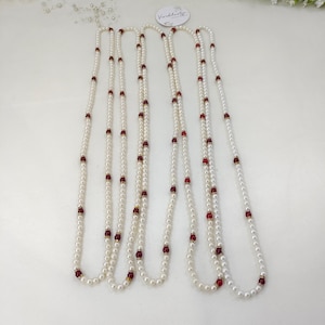 Puede incluir: Un collar largo de perlas blancas artificiales intercaladas con pequeñas cuentas redondas de color rojo intenso y espaciadores dorados. El collar está dispuesto en múltiples bucles, mostrando un patrón repetitivo de perlas blancas y acentos rojos.