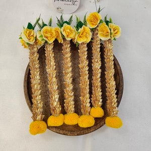 May include: Six golden flower garlands with yellow flowers and yellow pom-poms on a wooden plate. The garlands are made of woven fabric and have a traditional Thai design. The text "Vividcluster Unique as Rare" is on a white sticker.