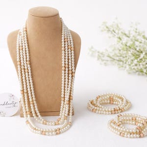 May include: A multi-strand necklace and two matching bracelets made of white faux pearls and small metallic gold beads. The necklace is displayed on a tan bust, and a circular white tag with the text Vividlustre is positioned to the left.