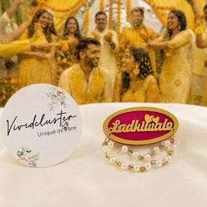 Puede incluir: Una tarjeta blanca circular con el texto Vividcluster Unique as are y detalles florales junto a un broche ovalado fucsia y dorado con el texto Ladkiwale, dos corazones peque&ntilde;os y dos hilos colgantes de perlas blancas y cuentas doradas.