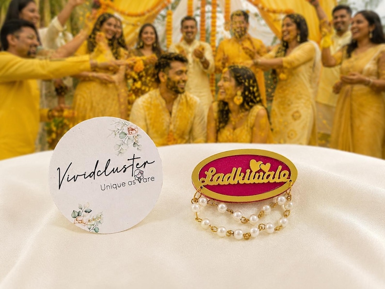 Puede incluir: Una tarjeta blanca circular con el texto Vividcluster Unique as are y detalles florales junto a un broche ovalado fucsia y dorado con el texto Ladkiwale, dos corazones peque&ntilde;os y dos hilos colgantes de perlas blancas y cuentas doradas.