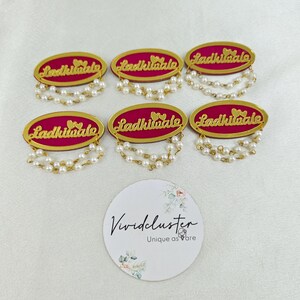 Puede incluir: Seis insignias ovaladas con fondo magenta y texto dorado que dice Ladkiwale junto a dos corazones peque&ntilde;os. Cada insignia est&aacute; adornada con dos hilos colgantes de perlas blancas artificiales en el borde inferior. Tambi&eacute;n se incluye una tarjeta circular blanca con el texto Vividcluster Unique as are.