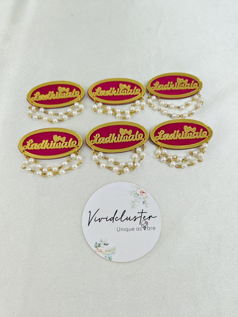 Puede incluir: Seis insignias ovaladas con fondo magenta y texto dorado que dice Ladkiwale junto a dos corazones peque&ntilde;os. Cada insignia est&aacute; adornada con dos hilos colgantes de perlas blancas artificiales en el borde inferior. Tambi&eacute;n se incluye una tarjeta circular blanca con el texto Vividcluster Unique as are.