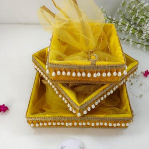 May include: Three-tiered yellow decorative trays with gold trim and white pearl accents. Each tray is square and lined with yellow fabric and a sheer yellow overlay. The trays are stacked, with a sheer yellow fabric draped over the top tray.