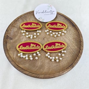 Puede incluir: Cuatro broches ovalados con fondo magenta, texto en cursiva dorada que dice Ladkiwale y dos peque&ntilde;os corazones dorados. Cada broche est&aacute; decorado con dos hilos colgantes de peque&ntilde;as perlas blancas artificiales en el borde inferior.