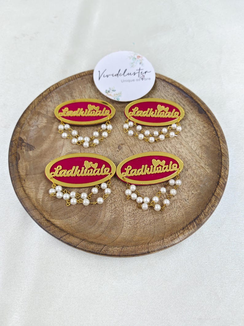 Puede incluir: Cuatro broches ovalados con fondo magenta, texto en cursiva dorada que dice Ladkiwale y dos peque&ntilde;os corazones dorados. Cada broche est&aacute; decorado con dos hilos colgantes de peque&ntilde;as perlas blancas artificiales en el borde inferior.