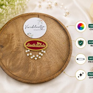 Puede incluir: Un broche de forma ovalada con un centro rojo intenso, el texto dorado 'Ladkiwale' y dos peque&ntilde;os corazones dorados. El broche est&aacute; adornado con dos hileras de perlas blancas artificiales y un marco de metal dorado con un cierre de seguridad.
