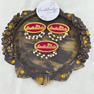 Puede incluir: Tres broches ovalados con centro magenta, bordes dorados y el texto "Ladkiwale" en letras doradas. Cada broche est&aacute; adornado con dos delicadas hileras de peque&ntilde;as perlas blancas artificiales que cuelgan del borde inferior.