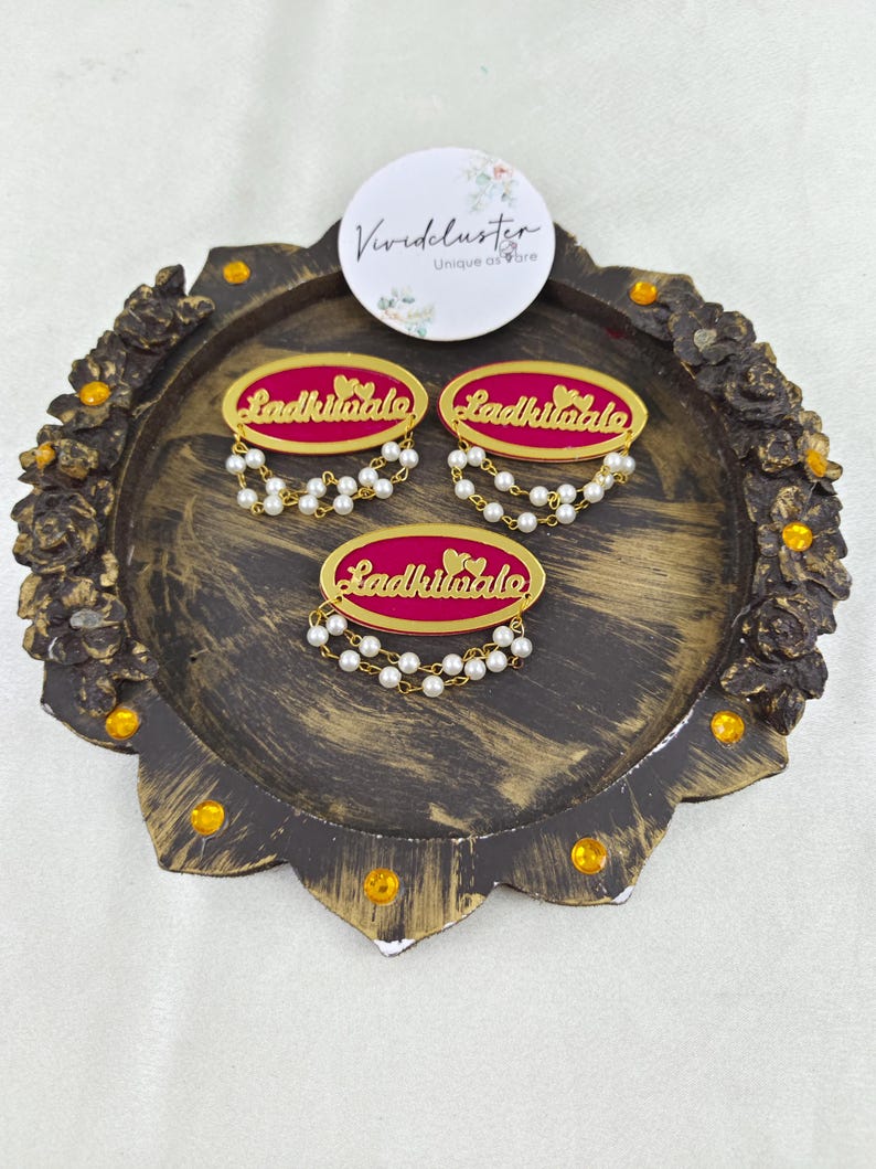 Puede incluir: Tres broches ovalados con centro magenta, bordes dorados y el texto "Ladkiwale" en letras doradas. Cada broche est&aacute; adornado con dos delicadas hileras de peque&ntilde;as perlas blancas artificiales que cuelgan del borde inferior.