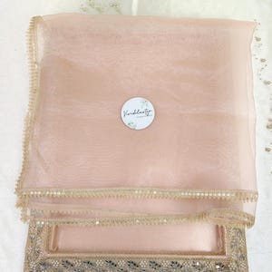 May include: A pale pink sheer scarf with a decorative, beaded edge. The scarf is folded and placed on a matching tray with an ornate, embroidered border. A small, round sticker with the text "Vividlustro" is visible.