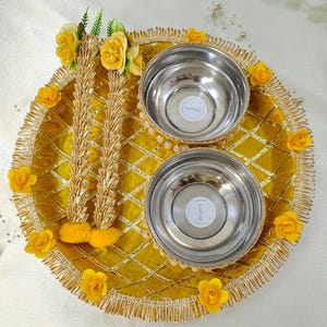 Puede incluir: Una bandeja dorada con forro de terciopelo amarillo y borde dorado. La bandeja tiene dos cuencos de plata y dos decoraciones de flores amarillas. La bandeja está decorada con un patrón de diamante.