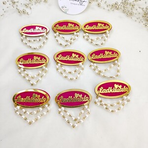 Puede incluir: Ocho broches de forma ovalada con un centro fucsia vibrante y un borde dorado. Cada broche muestra el texto Ladkiwale en letra dorada con dos peque&ntilde;os corazones encima, y dos delicadas cadenas de perlas blancas falsas colgando debajo.