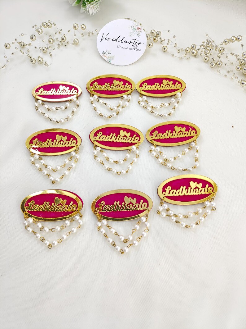 Puede incluir: Ocho broches de forma ovalada con un centro fucsia vibrante y un borde dorado. Cada broche muestra el texto Ladkiwale en letra dorada con dos peque&ntilde;os corazones encima, y dos delicadas cadenas de perlas blancas falsas colgando debajo.