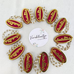 Puede incluir: Diez insignias ovaladas con centro magenta y borde dorado, cada una con el texto Ladkiwale y dos corazones peque&ntilde;os. Cada insignia est&aacute; unida a una doble hebra de perlas artificiales blancas. Una tarjeta blanca circular con el texto Vividcluster est&aacute; centrada.