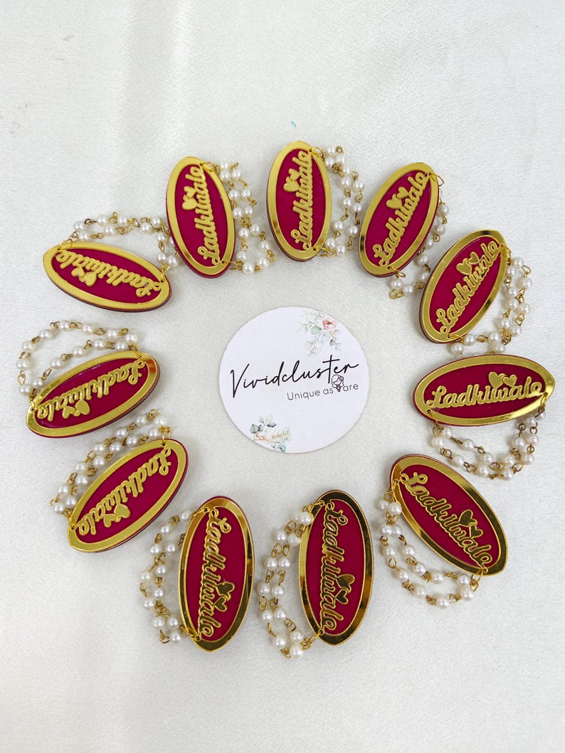 Puede incluir: Diez insignias ovaladas con centro magenta y borde dorado, cada una con el texto Ladkiwale y dos corazones peque&ntilde;os. Cada insignia est&aacute; unida a una doble hebra de perlas artificiales blancas. Una tarjeta blanca circular con el texto Vividcluster est&aacute; centrada.