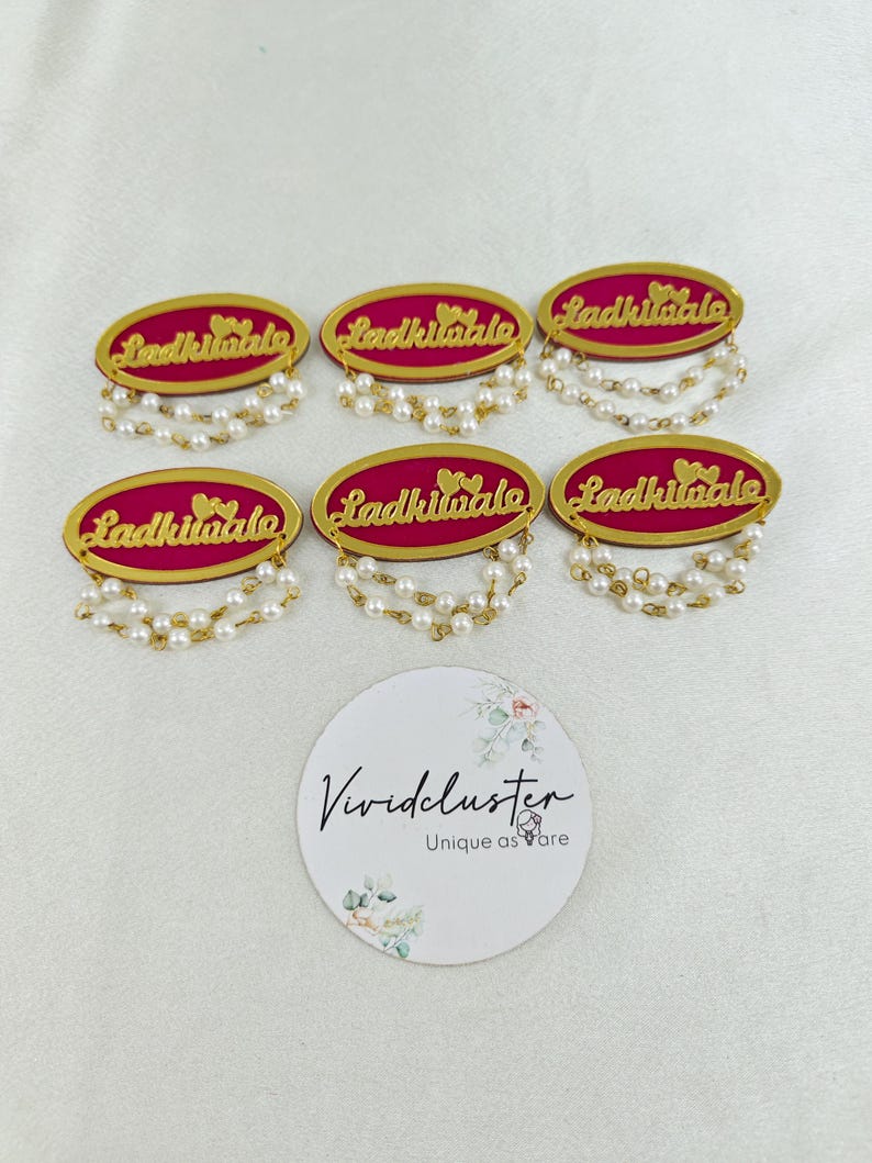 Puede incluir: Seis insignias ovaladas con un centro magenta, bordes dorados y el texto Ladkiwale en letra dorada. Cada insignia est&aacute; rematada con dos peque&ntilde;os corazones dorados e incluye dos hilos colgantes de perlas blancas artificiales en la parte inferior.