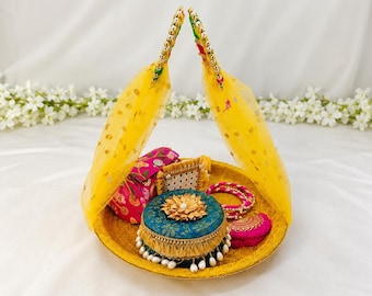 May include: A round golden tray features two arched handles draped with yellow polka-dot fabric. Inside, a teal circular box with a gold floral top, a pink floral pouch, a small rectangular beaded piece, and two pink bangles are arranged.