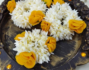 May include: Three circular floral garlands made of white jasmine-like blossoms and vibrant yellow fabric roses. These decorative wreaths are arranged on a dark, distressed circular wooden tray featuring gold-toned accents and embedded amber gems.