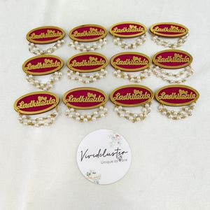 Puede incluir: Doce insignias ovaladas con un centro magenta, borde dorado y el texto Ladkiwale en letra dorada. Cada insignia est&aacute; adornada con dos hilos de peque&ntilde;as perlas blancas artificiales que cuelgan de la parte inferior. Una tarjeta blanca circular con el texto Vividcluster Unique as are se encuentra debajo de las insignias.