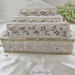 May include: A set of tiered white decorative boxes, each intricately decorated. The boxes feature silver mirrored details, faux pearls, and floral embroidery. The boxes are stacked, and a sheer white fabric drapes over the top. Measurements are in centimeters.
