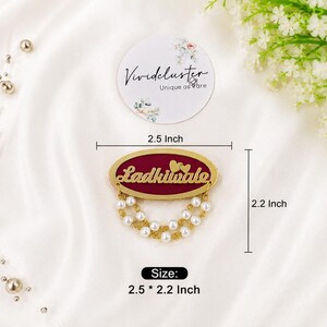 Puede incluir: Un broche de forma ovalada con un centro color granate profundo, el texto dorado 'Ladkiwale' y dos peque&ntilde;os corazones dorados encima. El broche est&aacute; adornado con dos hileras de perlas artificiales blancas y cadenas doradas, midiendo 6,35 por 5,59 cm.