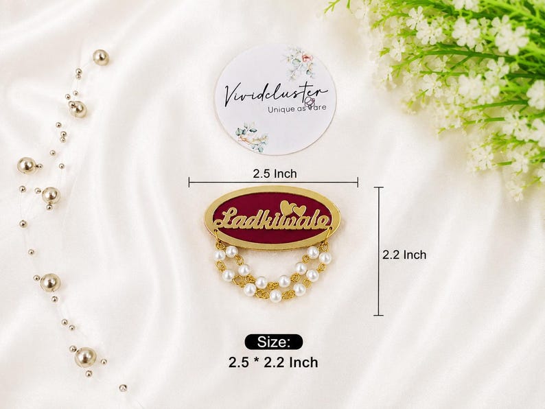 Puede incluir: Un broche de forma ovalada con un centro color granate profundo, el texto dorado 'Ladkiwale' y dos peque&ntilde;os corazones dorados encima. El broche est&aacute; adornado con dos hileras de perlas artificiales blancas y cadenas doradas, midiendo 6,35 por 5,59 cm.