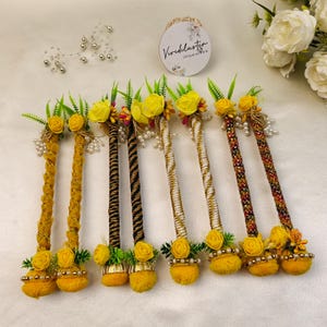 Op de afbeelding: Een verzameling decoratieve haaraccessoires, elk met een geel bloemendesign, groene bladeren en parelaccenten. De stelen zijn gewikkeld in gouden, zwarte en witte patronen. De accessoires zijn op een wit oppervlak geplaatst.
