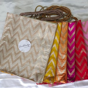 Puede incluir: Un conjunto de siete bolsas de tela con un patrón de chevron. Las bolsas son de varios colores, incluyendo dorado, amarillo, naranja, rosa, rojo y magenta. Las bolsas tienen un hilo dorado tejido en la tela. Las bolsas tienen una etiqueta que dice "Vividlustro Design Store".