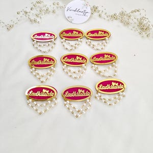 Puede incluir: Ocho insignias ovaladas con un centro rosa brillante, borde dorado y el texto "Ladkiwale" en letra dorada. Cada insignia est&aacute; adornada con dos hilos colgantes de peque&ntilde;as perlas artificiales blancas fijadas en el borde inferior.