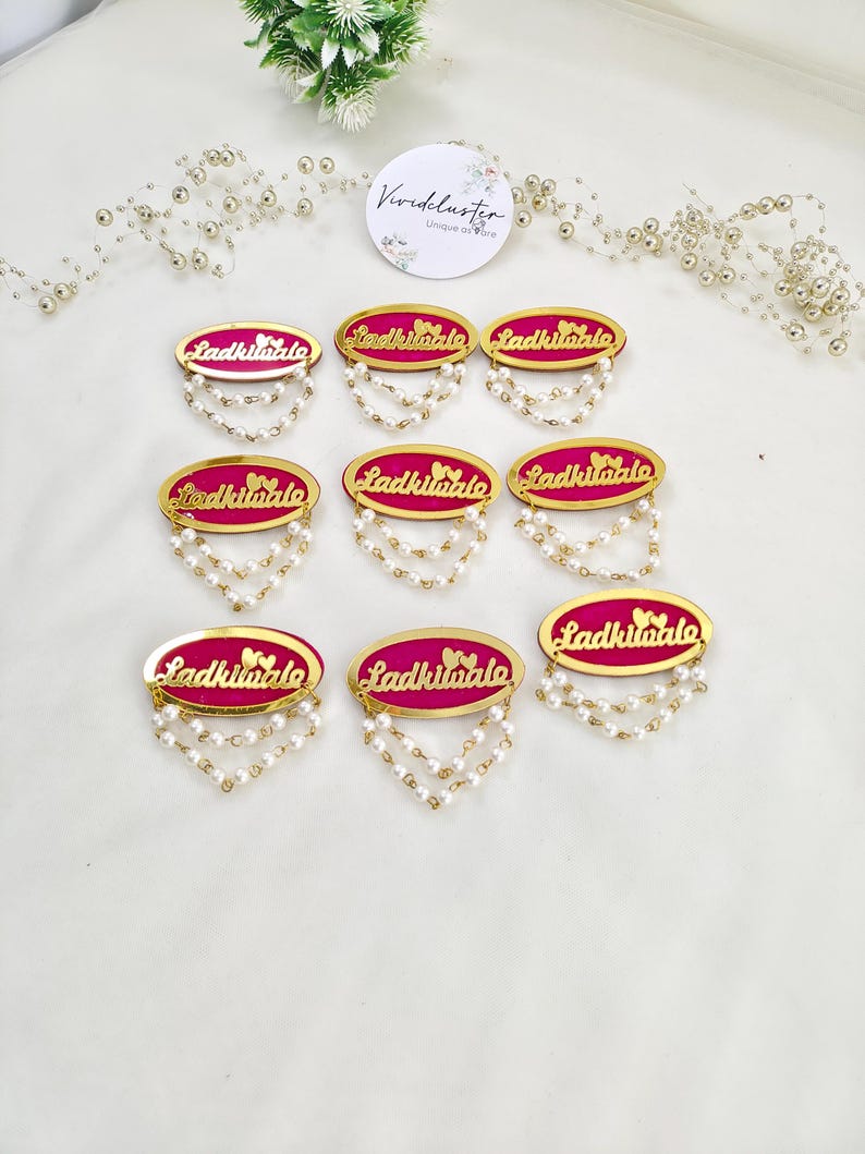 Puede incluir: Ocho insignias ovaladas con un centro rosa brillante, borde dorado y el texto "Ladkiwale" en letra dorada. Cada insignia est&aacute; adornada con dos hilos colgantes de peque&ntilde;as perlas artificiales blancas fijadas en el borde inferior.