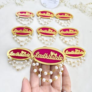 Puede incluir: Broches de forma ovalada con un centro rosa brillante, bordes dorados y la inscripci&oacute;n Ladkiwale. Cada pieza est&aacute; decorada con dos hilos colgantes de perlas blancas artificiales sujetos al borde inferior del marco dorado.