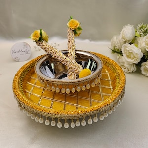 May include: A decorative yellow fabric tray with a gold lattice pattern and hanging white faux pearl beads. A silver metal bowl sits in the centre, adorned with two gold-toned floral garlands featuring yellow roses and a small yellow pom-pom.