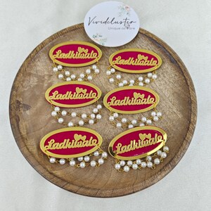 Puede incluir: Seis insignias ovaladas con un fondo magenta vibrante, texto en cursiva de color dorado que dice Ladkiwale y dos corazones peque&ntilde;os. Cada insignia est&aacute; decorada con dos hilos colgantes de peque&ntilde;as perlas blancas artificiales.