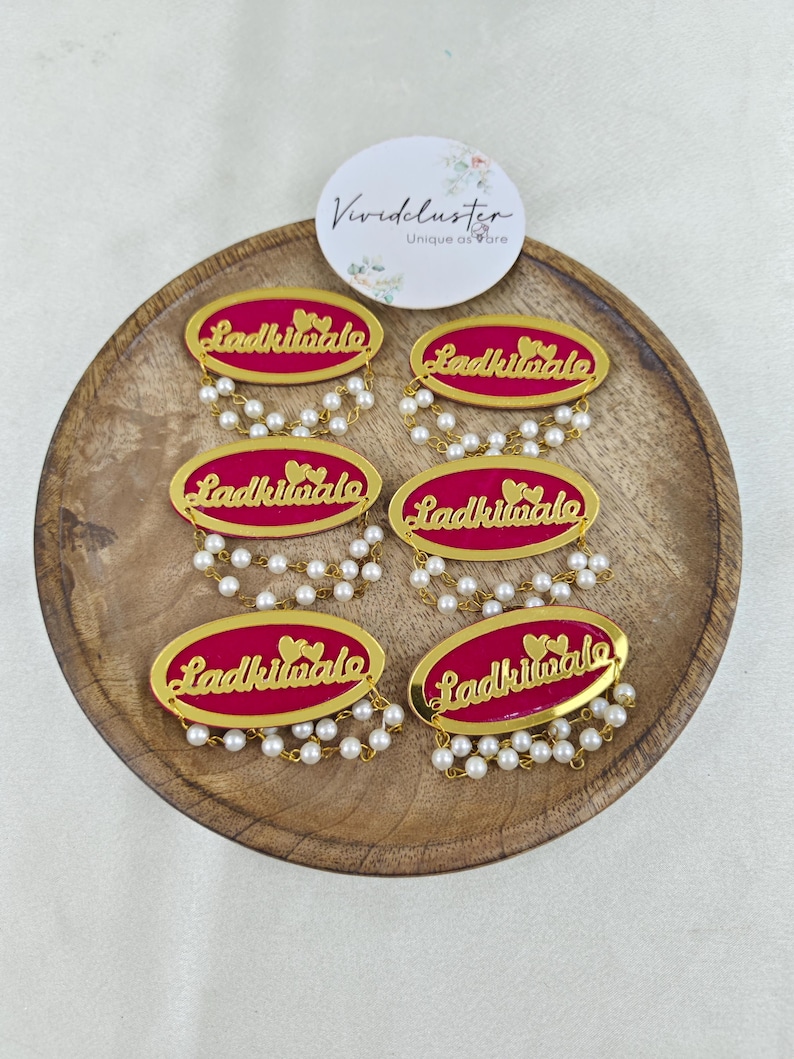 Puede incluir: Seis insignias ovaladas con un fondo magenta vibrante, texto en cursiva de color dorado que dice Ladkiwale y dos corazones peque&ntilde;os. Cada insignia est&aacute; decorada con dos hilos colgantes de peque&ntilde;as perlas blancas artificiales.