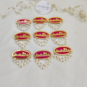 Puede incluir: Ocho insignias ovaladas con un centro rosa brillante, bordes dorados y el texto Ladkiwale. Cada insignia est&aacute; decorada con dos peque&ntilde;os corazones dorados y dos hilos colgantes de perlas artificiales blancas en el borde inferior.