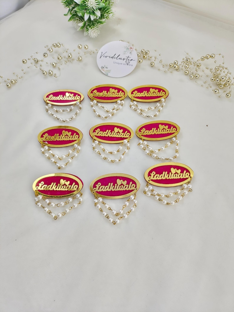 Puede incluir: Ocho insignias ovaladas con un centro rosa brillante, bordes dorados y el texto Ladkiwale. Cada insignia est&aacute; decorada con dos peque&ntilde;os corazones dorados y dos hilos colgantes de perlas artificiales blancas en el borde inferior.
