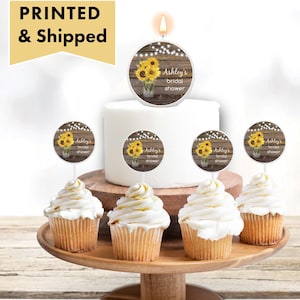 May include: A white cake with a sunflower and woodgrain design topper that says "Ashley's bridal shower". There are six cupcakes with sunflower and woodgrain design toppers that say "Ashley's bridal shower".
