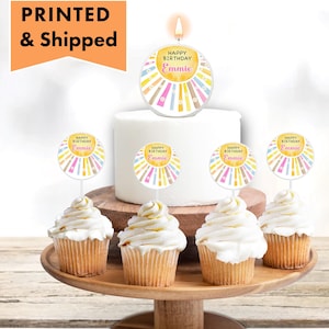 May include: A set of six yellow, pink, and blue cupcake toppers with the text "Happy Birthday Emme" on each. The toppers are on a white cake and cupcakes.