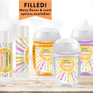 May include: Four hand sanitizer bottles with labels that say "Happy Birthday Emmie" and a sunburst design. The bottles are filled with clear liquid and have white lids. There are also two lip balm tubes with the same design and text.