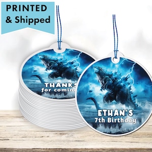 May include: Two round, white, printed favor tags with blue string. The tag on the left says "Thanks for coming". The tag on the right has a blue and white image of a Godzilla-like monster and says "Ethan's 7th Birthday".