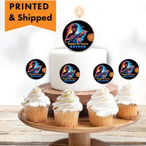 May include: A white cake with a blue and orange gamer themed candle on top. Six cupcakes with blue and orange gamer themed toppers are arranged around the cake on a wooden tray. The cake and cupcakes are decorated with the text "Happy Birthday Hayden".