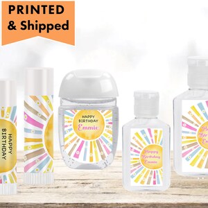 May include: Set of four personalized hand sanitizer bottles and lip balm tubes with a yellow sun and rainbow design. The labels say "Happy Birthday Emmie".